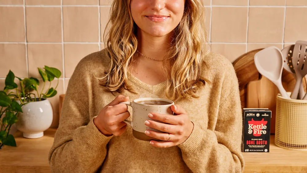 A woman holding a warm mug in her kitchen, a Kettle & Fire carton beside her